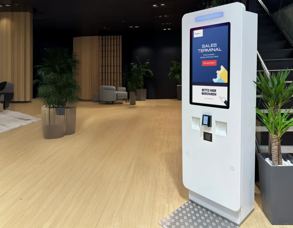 A modern self-service sales terminal with a large touchscreen stands in a stylish indoor space. The interface prompts users to interact, with integrated payment and scanning options.